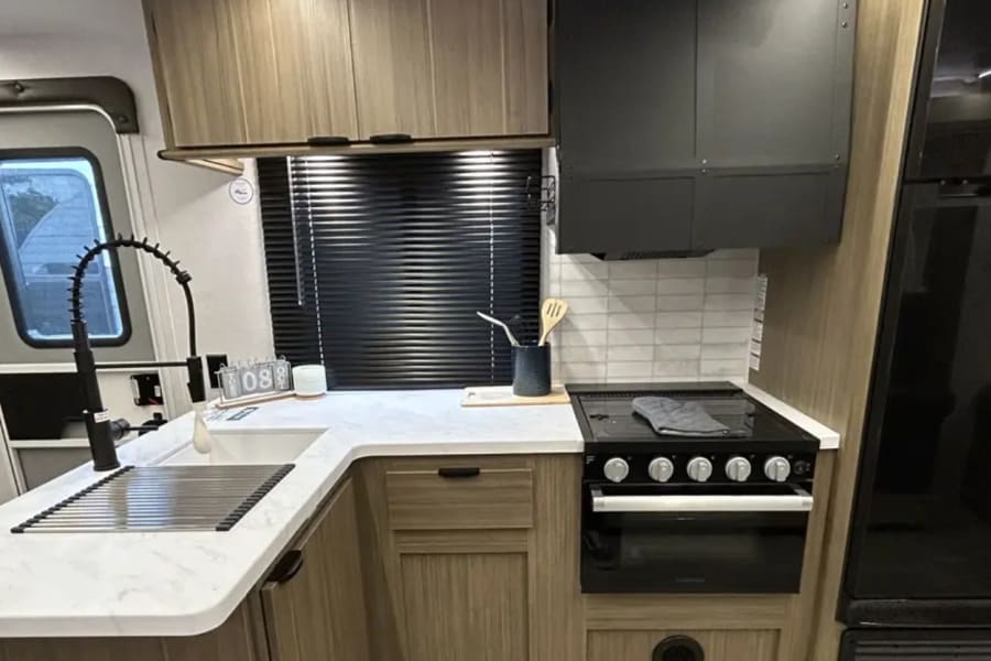 RV rental kitchen with a modern design: black stove, oven, sink, and refrigerator. Features light wood cabinets and white countertops.