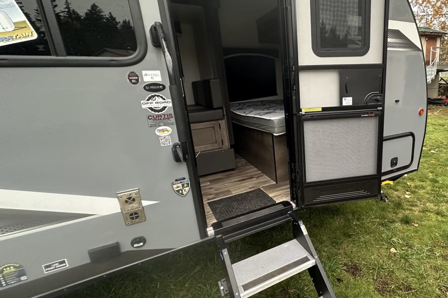 An RV rental with its side door open, revealing a cozy interior with a bed. Extended steps lead to the grassy ground.