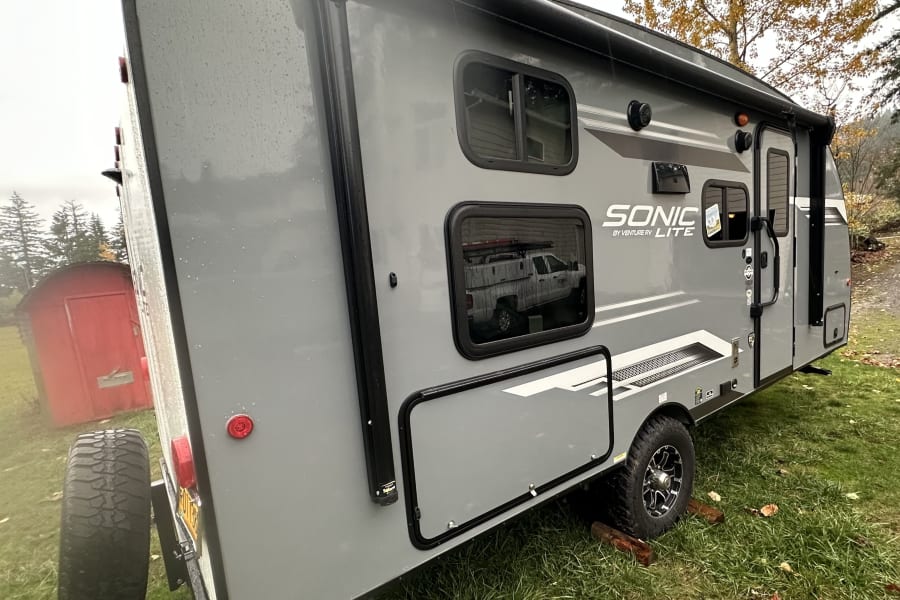 Gray Sonic travel trailer for your next RV rental trip, parked on wet grass with trees and a red shed in background.