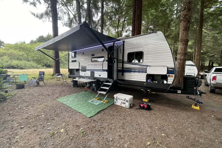 Enjoy this spacious RV rental campsite featuring an extended awning, outdoor seating, and a lush forest backdrop.
