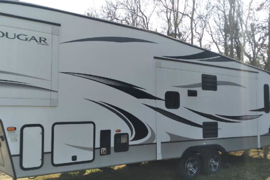 White Cougar fifth-wheel RV rental, featuring black and gray graphics and multiple slide-outs, parked outdoors.