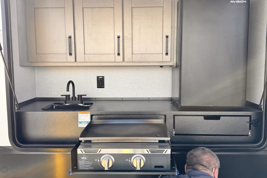 RV rental outdoor kitchen setup with sink, fridge, cabinets, and a Capital Griddle, being accessed by a person.