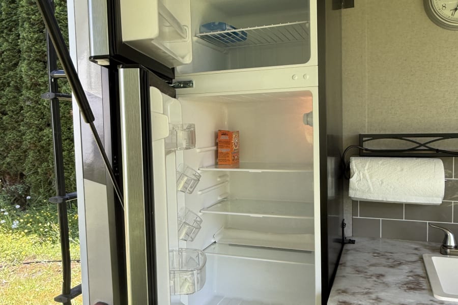 An open RV rental refrigerator in an outdoor kitchen area with a counter, sink, and paper towels, next to an exterior ladder.