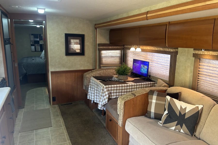 Cozy RV rental interior showcasing a warm living/dining area with a TV, adjoining bedroom, and a peek of the kitchen.