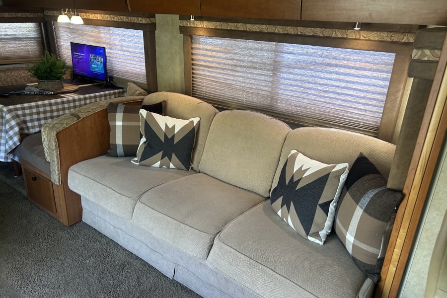 Cozy RV rental interior with a light-colored sofa, patterned pillows, and a dining area with a TV and plant.