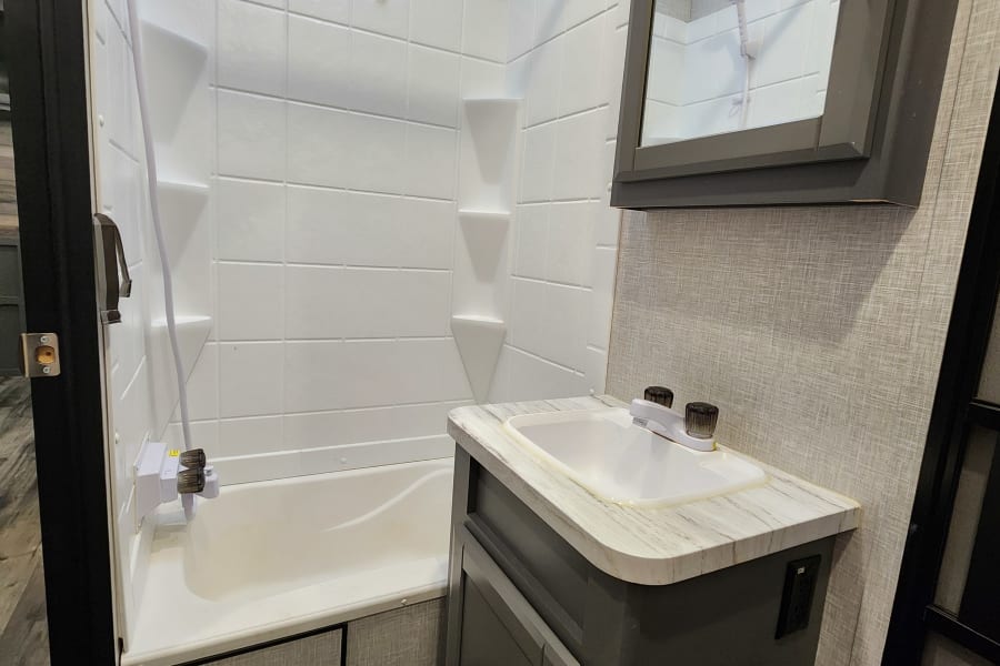 RV rental bathroom: white shower-tub, handheld shower, dark vanity with sink, mirror. Practical and clean space.