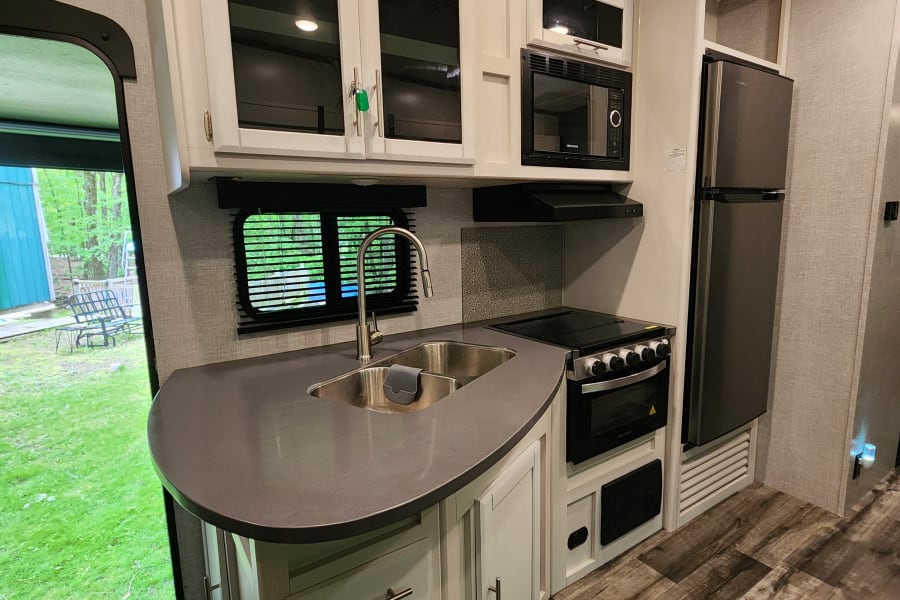 Modern RV rental kitchen: white cabinets, dark counters, sink, and appliances. Bright interior with view to green yard.