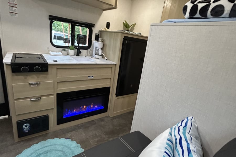 Cozy RV rental interior featuring a kitchen with stove/sink, electric fireplace, elevated bed, and comfy sofa.