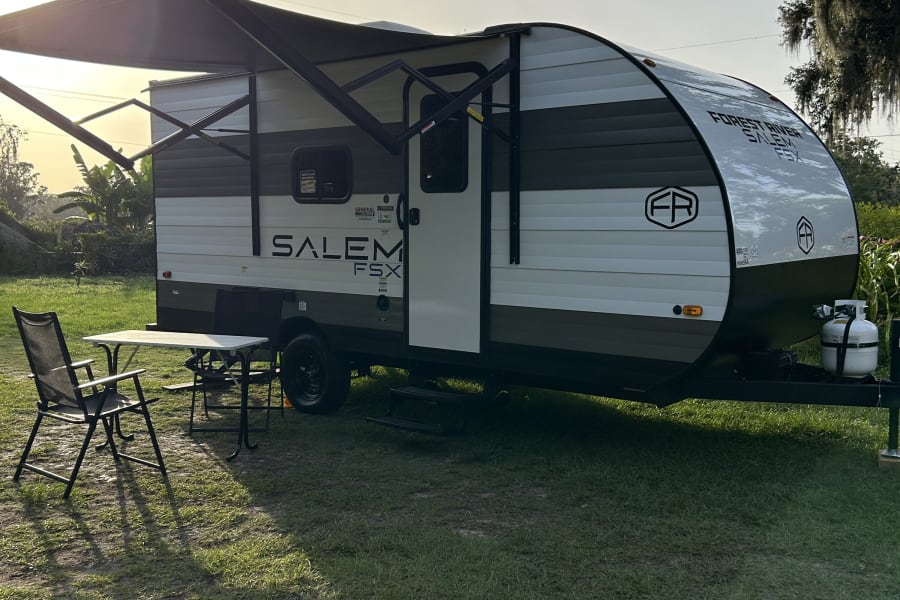 Sunlit RV rental: Forest River Salem FSX travel trailer with extended awning, table, chairs on grass, Spanish moss trees visible.