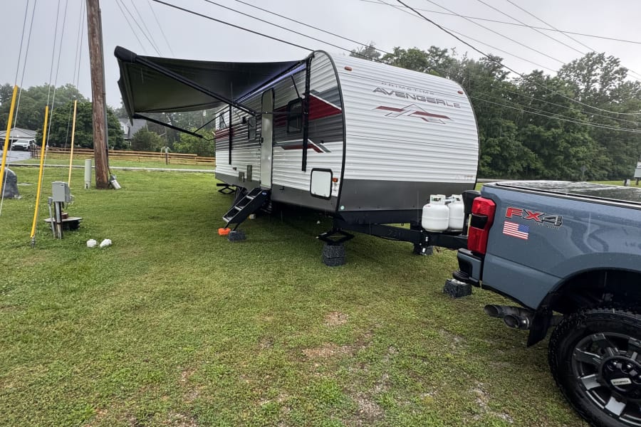 An RV rental (Prime Time Avenger travel trailer) is parked with its awning extended, hitched to a grey Ford truck at a campsite.