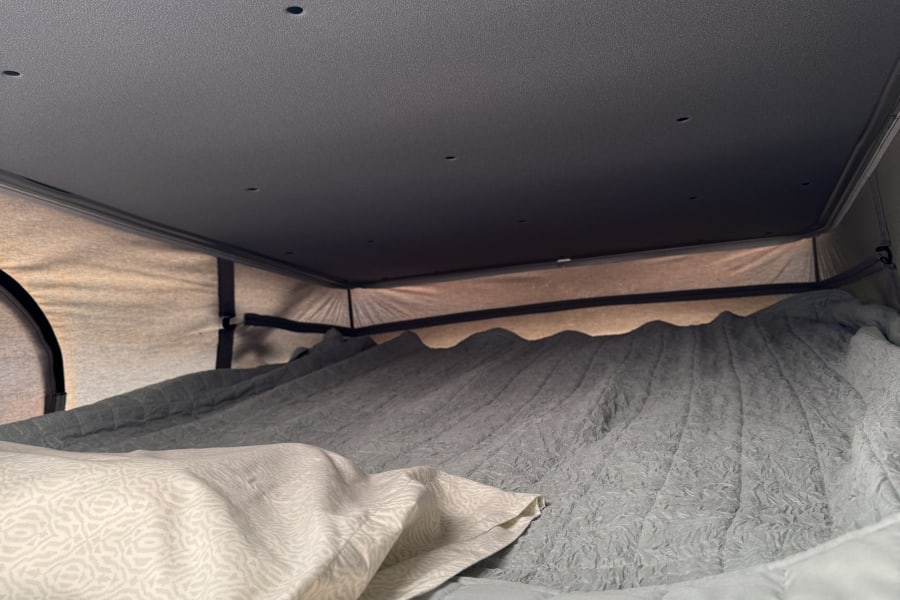 Inside an RV rental: a cozy bed with a patterned pillow, quilted blanket, and textured bedding under a ceiling with small lights.