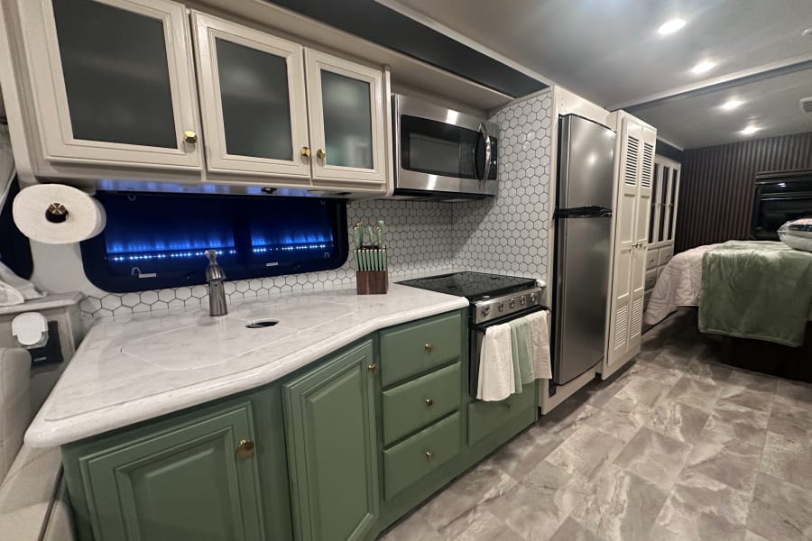 Modern RV rental kitchen with green/white cabinets, stainless steel appliances, hexagon tile backsplash, and a bedroom visible.