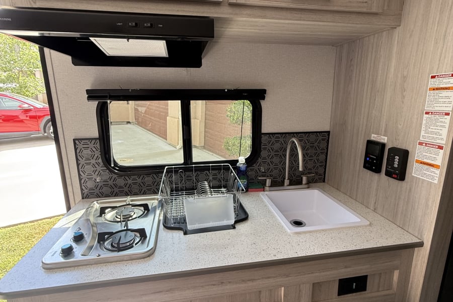 RV rental kitchen: Features light wood cabinetry, a two-burner stove, sink, and electric fireplace/heater below.