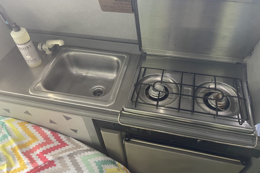Compact RV rental kitchen with sink, faucet, dish soap, and two-burner stove. Ideal for on-the-go cooking.