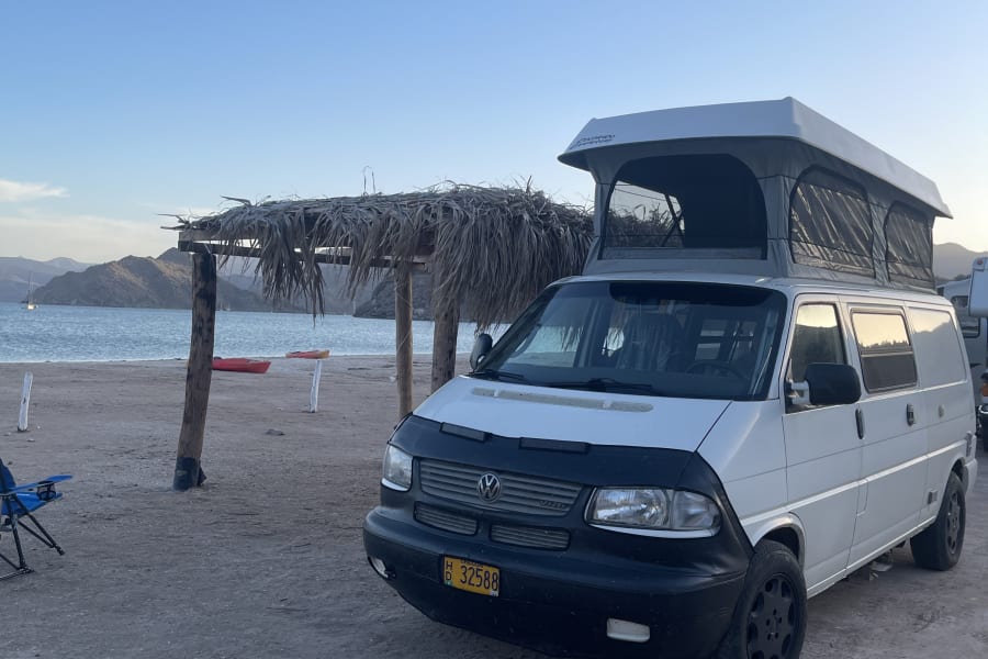Beach camping with an RV rental: a white VW camper van with a pop-top roof by the ocean, mountains in the background.
