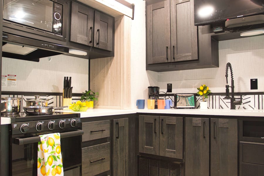 A modern RV rental kitchen featuring dark wood cabinets, white countertops, a stove, microwave, and a black faucet at the sink.