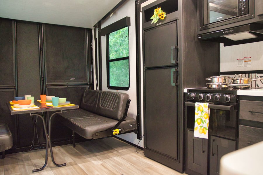 RV rental interior: A compact, modern kitchen with fridge, oven, microwave, and dining area with bench seating and colorful dishes.