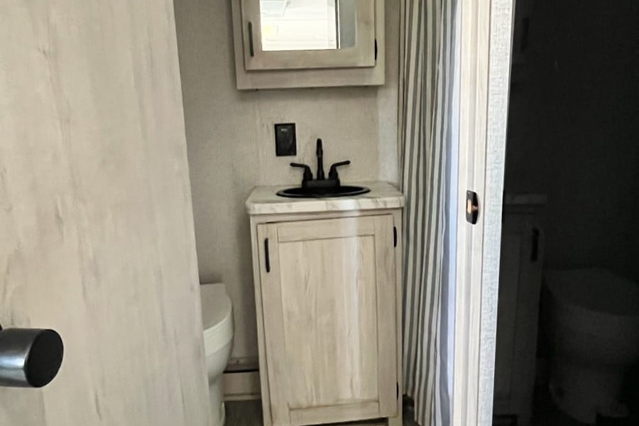 Modern RV rental bathroom: light wood vanity, dark sink, mirrored cabinet, toilet, and striped shower curtain.