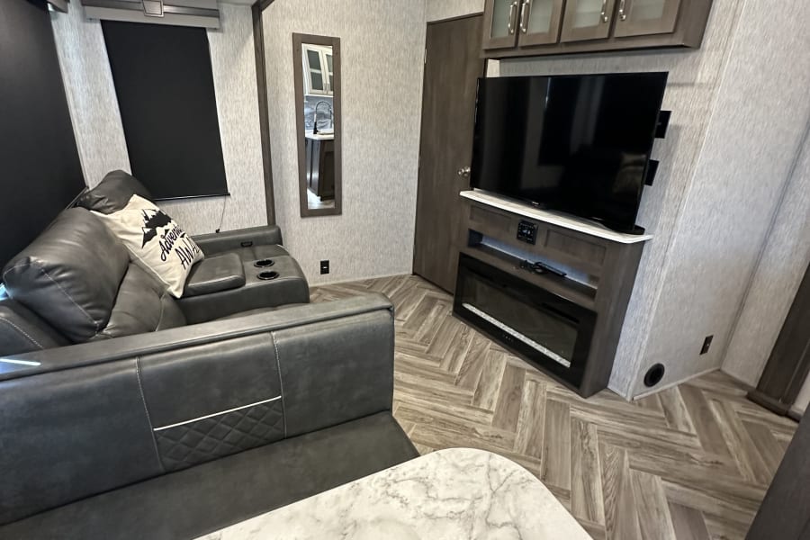 Cozy RV rental living space with modern grey recliners, TV, fireplace, and herringbone floor. Marble counter in foreground.