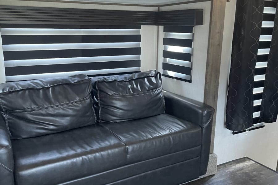 This RV rental offers a modern interior with a dark faux leather sofa, striped window blinds, and elegant ceiling lighting.