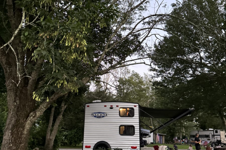Dusk at a leafy campground, showing a family near their white Salem travel trailer rv rental under tall trees.