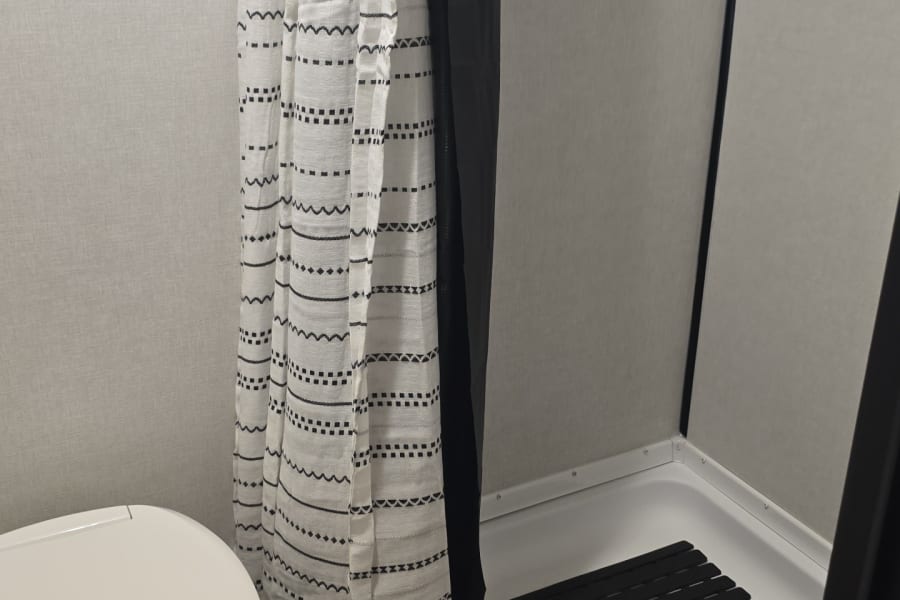 Compact RV rental bathroom: features a white toilet, patterned shower curtain, and shower stall with a black mat.