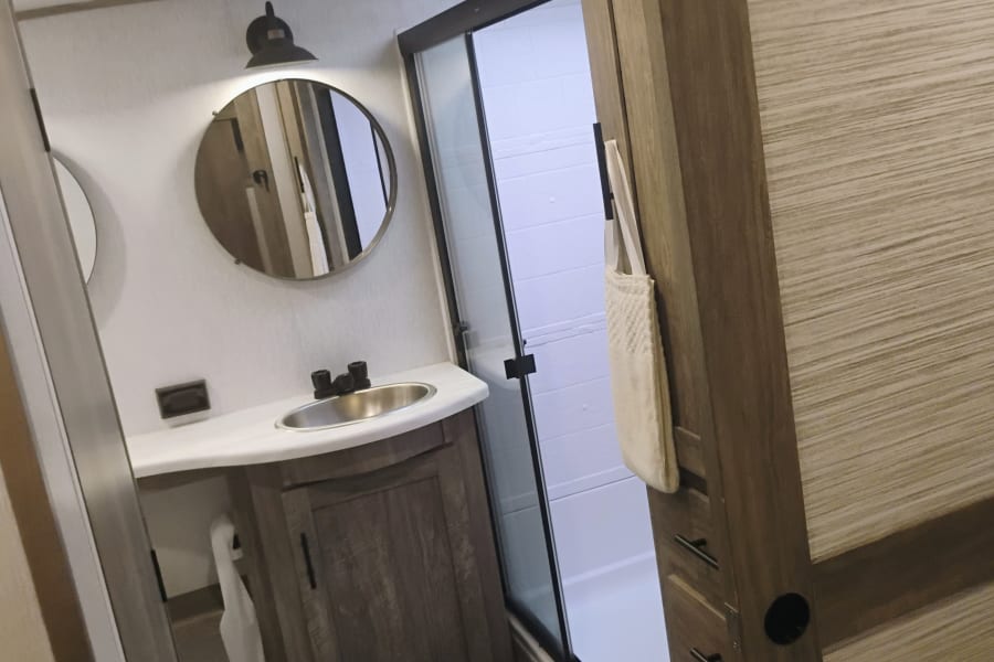 Modern RV rental bathroom with a vanity sink, round mirror, and clear glass shower, ideal for travel.
