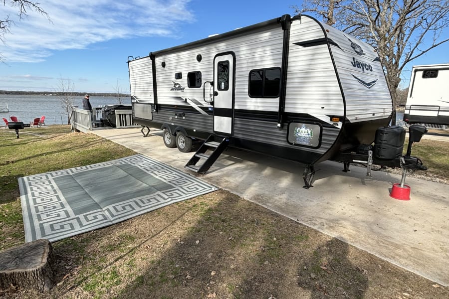 Spacious Jayco RV rental at a scenic lakefront campsite. Man on deck, outdoor rug beside the camper for lakeside camping.