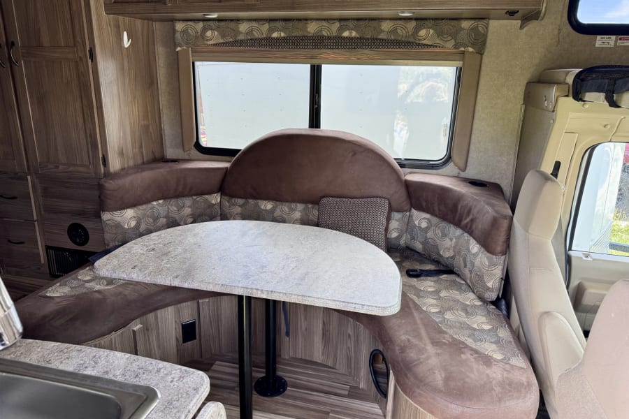 Spacious RV rental dinette with U-shaped booth seating, table, wood-grain cabinetry, and cockpit view.