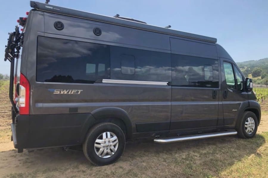 Dark gray Swift rv rental camper van with bike rack & awning, parked outdoors in a sunny vineyard.