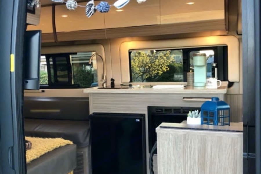 View inside a clean RV rental: compact kitchen, seating/bed, and bright interior lights. Perfect for road trips.