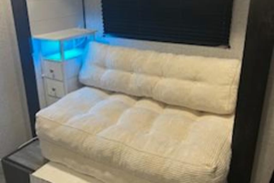 Compact RV rental space with a futon sofa over storage, illuminated side table, and a black window blind.