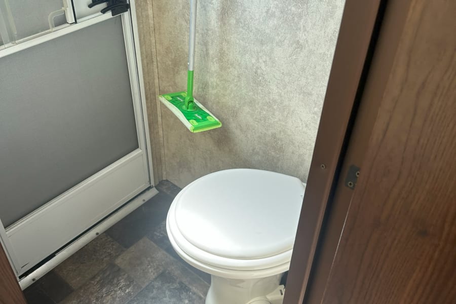 This RV rental features a compact bathroom with a white toilet, screen door, and wall-mounted Swiffer mop.