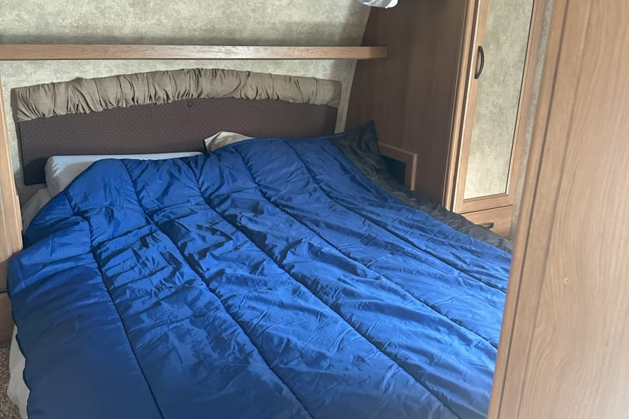 Cozy RV rental bedroom: blue comforter on bed, wood-toned overhead cabinets, and built-in mirrored storage.