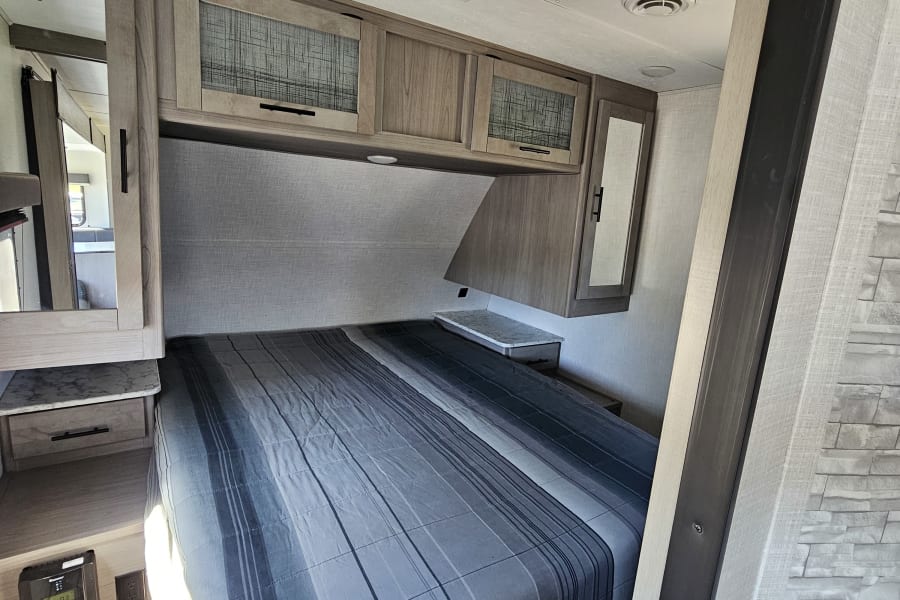 Inside an RV rental: a compact bedroom featuring a comfortable striped bed, overhead cabinets, and power outlets.