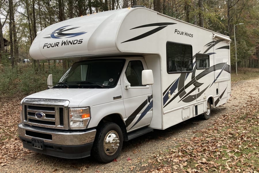 A white 'Four Winds' Class C rv rental with graphic decals parked on a leaf-covered path in a wooded campground.