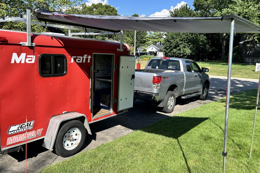 Motorsport trailer with open side, deployed awning, hitched to a silver pickup. Explore options for RV rental adventures.