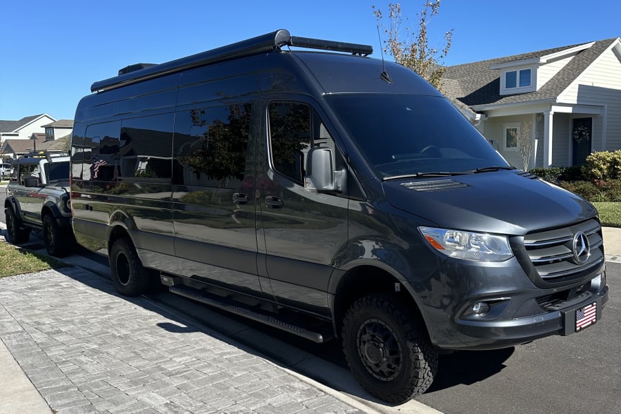 Dark grey Mercedes-Benz Sprinter camper van, perfect for an RV rental, parked on a sunny street next to a silver Ford Bronco.