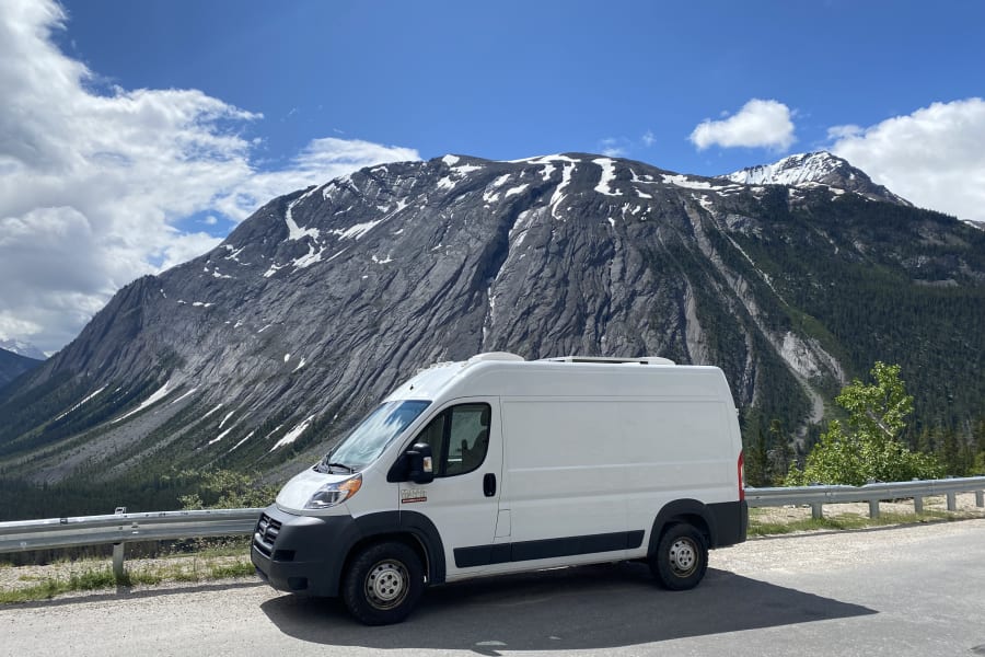 An RV rental (Ram Promaster) parked on a mountain road with towering, snow-dusted peaks and a blue sky.