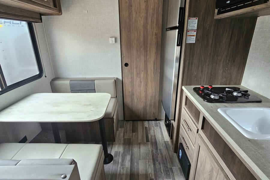 View inside an rv rental, showing a compact kitchen with sink, stove, microwave, and refrigerator, opposite a bright dinette.
