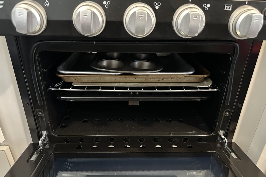 Inside an RV rental oven, showing metal baking pans on a rack. Control knobs for range and oven visible above the open door.