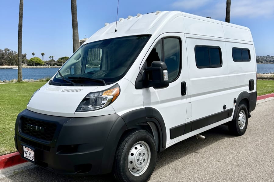 White Ram ProMaster camper van, an RV rental, parked by a sunny California waterfront with palm trees.