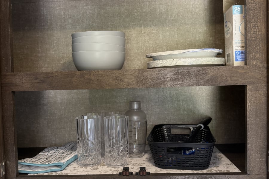 Inside an RV rental: a neatly stocked kitchen cabinet with bowls, paper plates, cutlery, drinking glasses, and a cocktail shaker.
