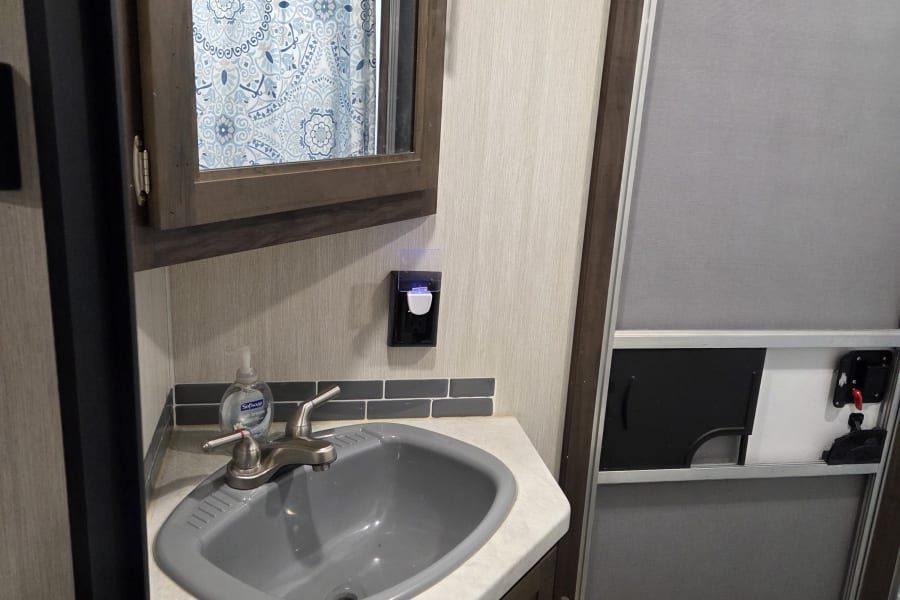 RV rental interior: A compact, clean bathroom with a gray sink, vanity, mirror, and blue patterned shower curtain.
