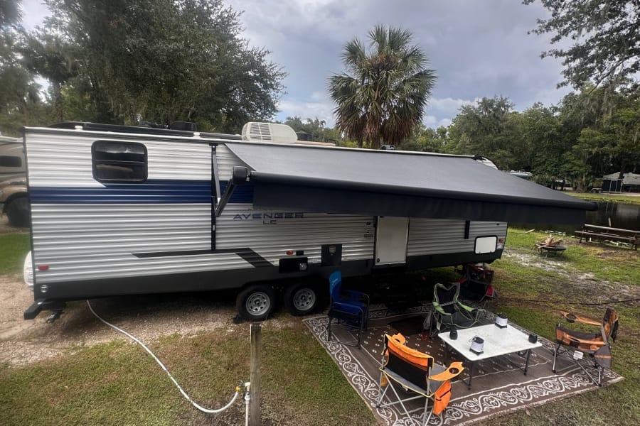 RV rental camping setup: travel trailer with extended awning, outdoor rug, table, and chairs under cloudy skies.