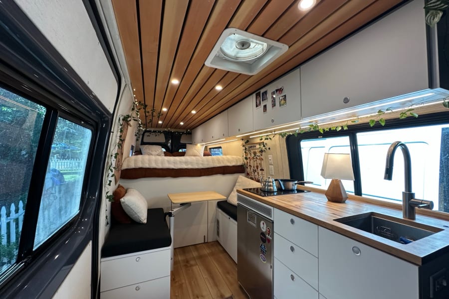 Step inside this modern RV rental: a spacious camper van with a cozy bed, kitchen, seating, and wooden ceiling.