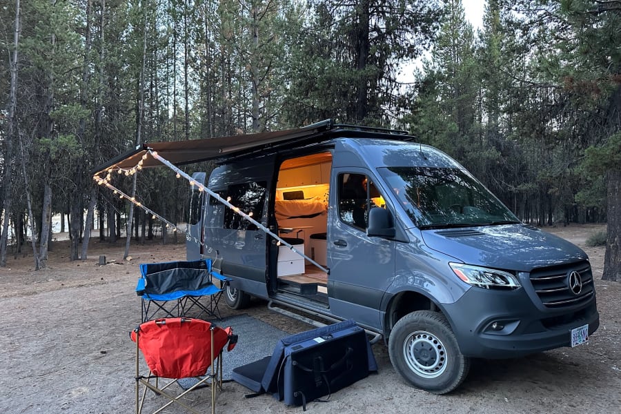 Cozy Mercedes Sprinter RV rental set up for camping in a pine forest, with string lights and chairs outside. Perfect for van life.
