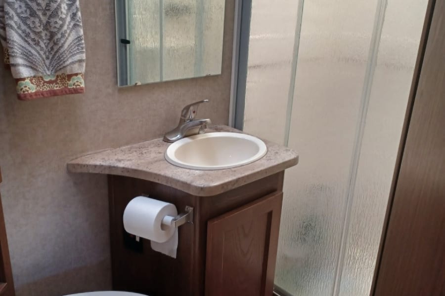 Compact RV rental bathroom, featuring a toilet, corner sink with mirror, and a frosted shower stall.