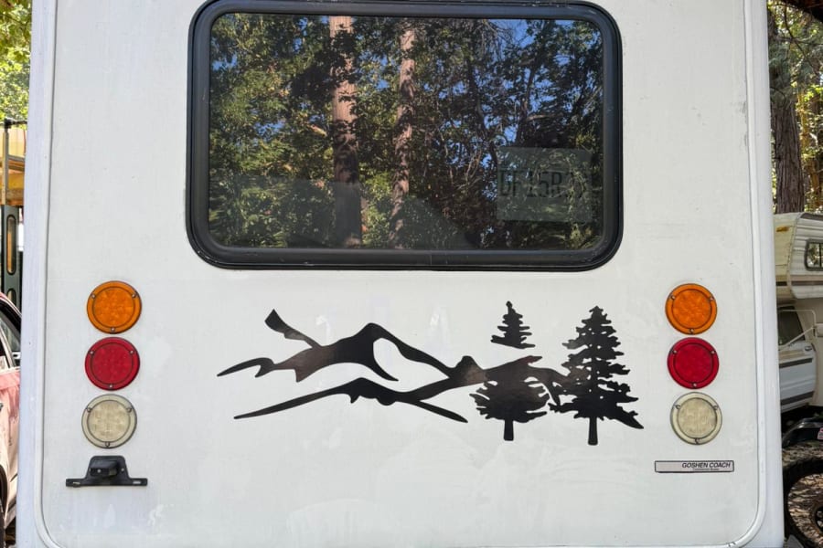 Rear of a white Goshen Coach RV, decorated with mountain & tree decals. Great for your next outdoor RV rental adventure!