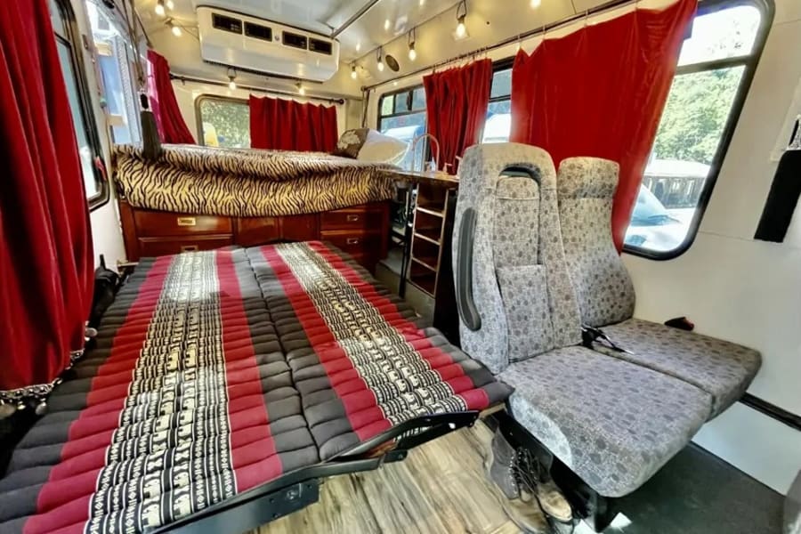 Inside an RV rental: elevated zebra-print bed, red curtains, a floor mat, and passenger seats for comfy travel.